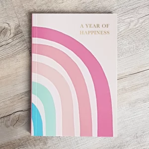 This happiness journal is laid on a surface showing the front cover. Multicoloured rainbow adorns it and gold foil writing A year of Happiness